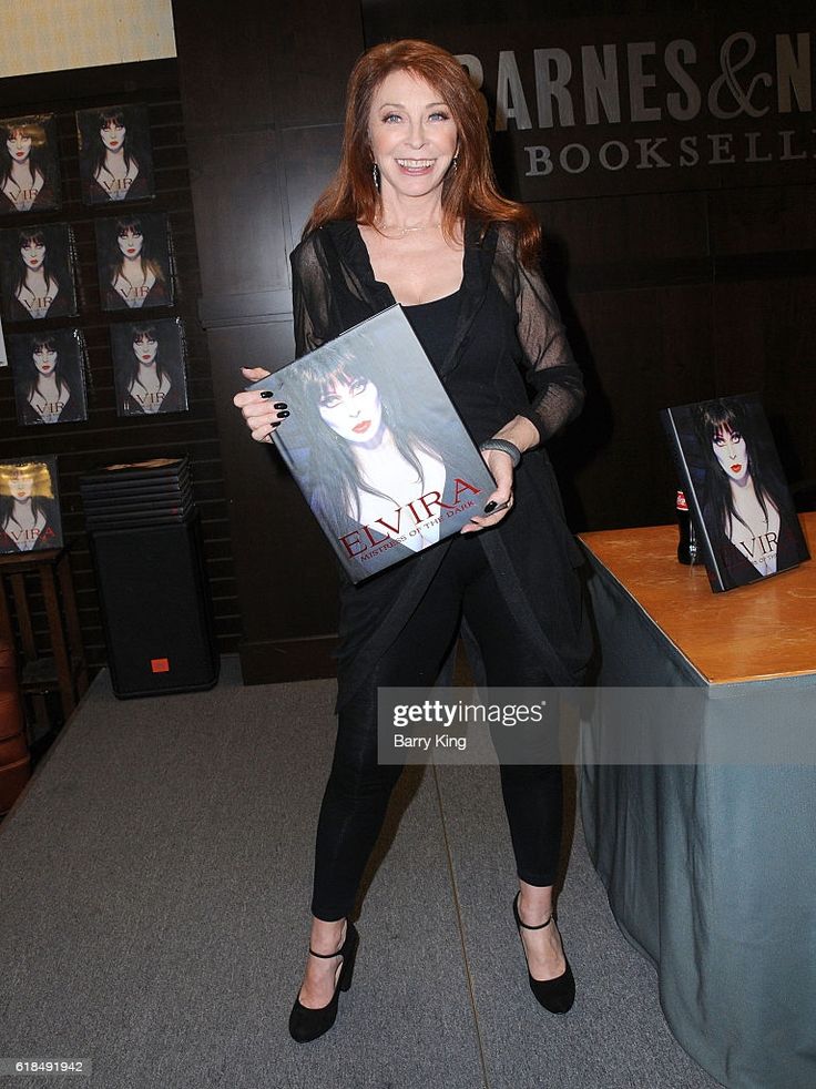 elvira, mistress of the dark, is signing her book, "elvira, mistress of the dark", for a fan at a barnes & noble bookstore.  she is dressed in black and wearing high heels. she is smiling and holding a book in her hands. the fan is standing behind her.  the book cover features a photo of elvira in her iconic black dress and wig.  a stack of books sits on the table, and a sign for a barnes & noble book signing event.