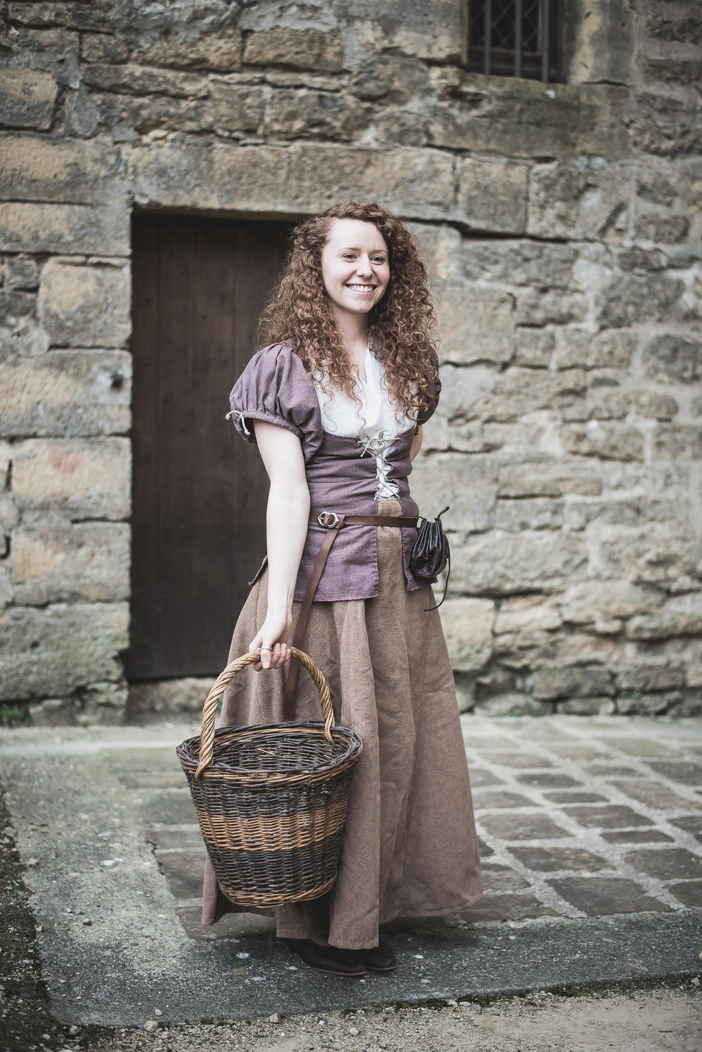 a full shot depicts a woman in a period costume, standing outdoors with a basket in hand. she has curly brown hair and is smiling. the woman is wearing a peasantstyle dress consisting of a light purple corsetlike top over a white blouse, cinched at the waist with a brown belt, and a long brown skirt. a small black pouch hangs from her belt. she's holding a brown and black woven basket in her left hand. the background features a stone building with a doorway and a small barred window. the ground is paved with cobblestones.