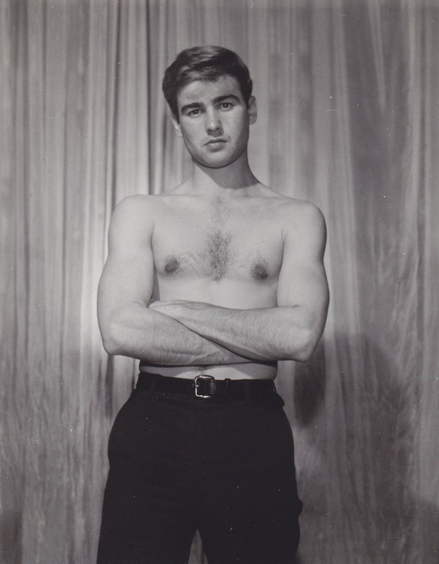 a black and white portrait of a young man with his arms crossed, standing confidently against a draped backdrop. he is shirtless, wearing dark pants, and has a serious expression on his face. the studio lighting highlights his physique and creates a sense of depth.