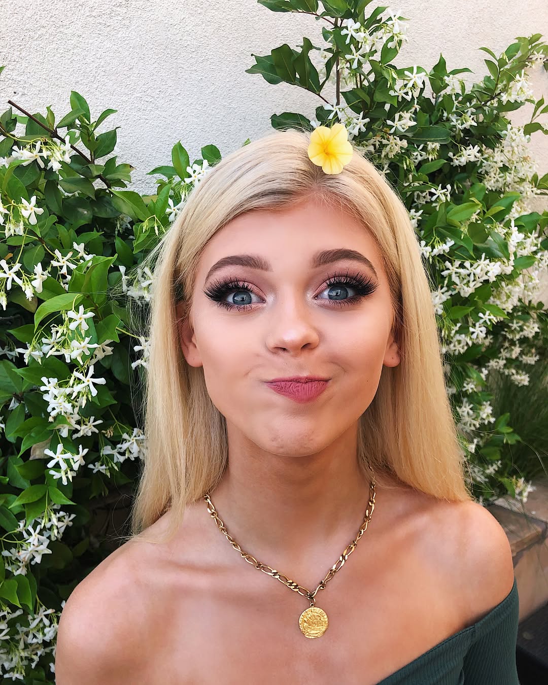 a young woman with blonde hair is posing in front of a wall covered in greenery and white flowers. she is wearing a gold necklace with a coin pendant and an offtheshoulder green top. she has a yellow flower in her hair, and she is pouting her lips. she is wearing makeup including defined eyelashes.