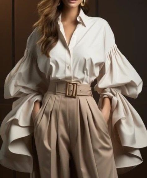 a woman stands posing in a stylish and elegant outfit. she is wearing a white blouse with dramatic, voluminous sleeves and a pair of highwaisted beige pants cinched with a gold buckle belt.  her hair is styled in loose waves and she is accessorized with gold earrings. the background is a neutral, dark brown color, allowing the focus to remain on the woman and her outfit. the image evokes a sense of sophistication and confidence.