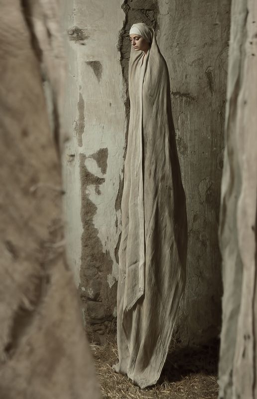 a woman stands draped in a long, neutralcolored cloth within a rustic, stonewalled interior. she wears a head covering and stands on a bed of hay. the atmosphere is aged and contemplative, with the textures of the stone and cloth emphasizing the traditional, modest setting.