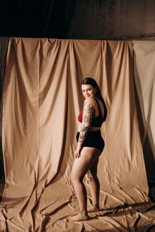 a plussize woman with tattoos on her arms is confidently posing in a burgundy bra and black shorts against a beige fabric backdrop. she's smiling slightly and looking over her shoulder at the camera, showcasing body positivity and selfacceptance. the lighting is soft and the overall mood is one of empowerment and celebrating body diversity.