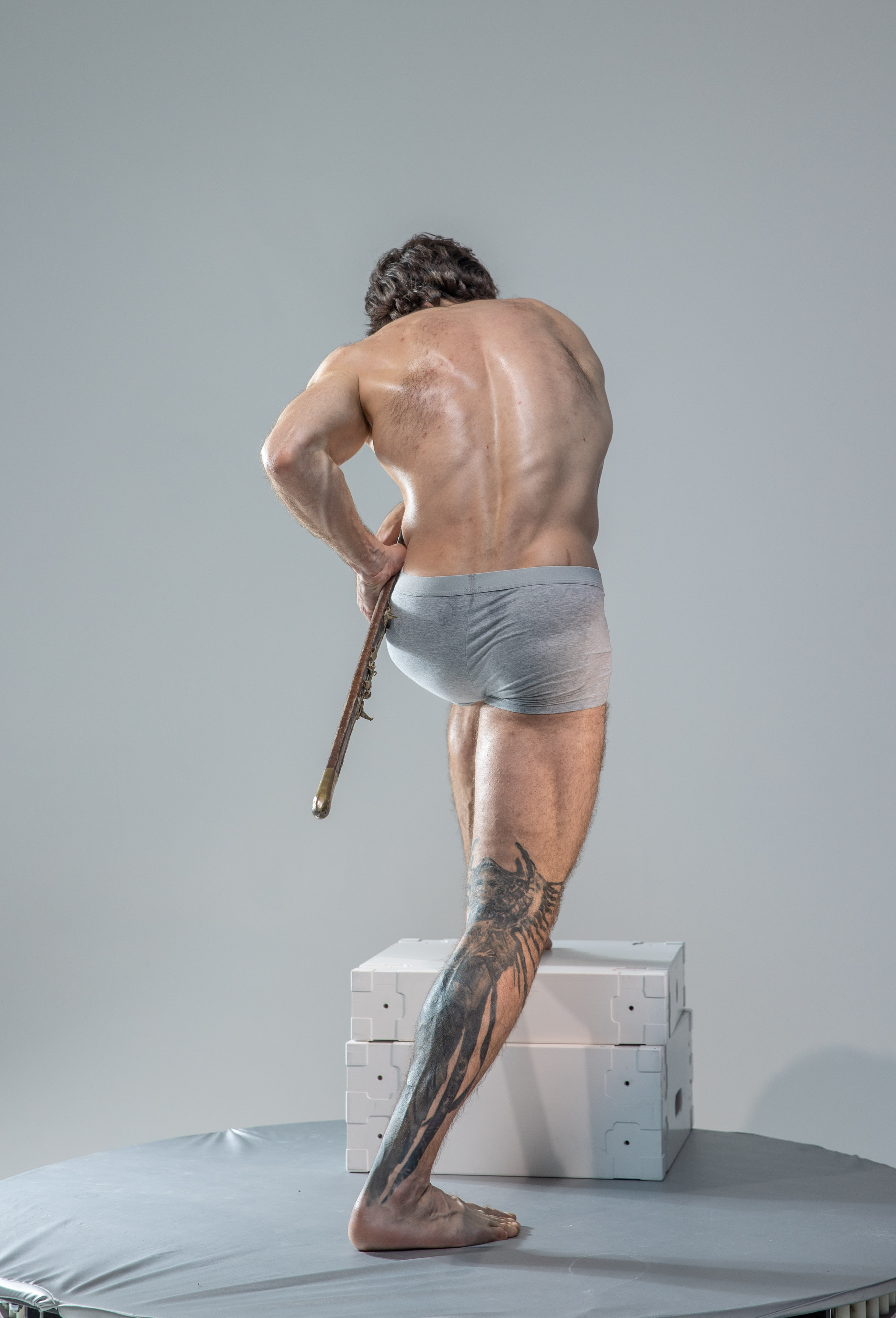 a muscular man with tattoos stands with his back to the camera in a classic contrapposto pose. he holds a long, thin object, possibly a pointer stick, in his right hand. the man is shirtless and wears only gray briefs. he is positioned on a gray platform with his left foot raised on two white blocks. the background is a solid light gray, suggesting a studio setting. the lighting is soft and even, illuminating the subject's form without harsh shadows.