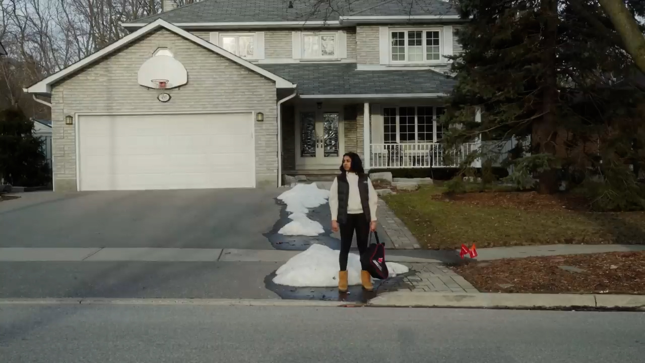a woman stands at the end of a driveway in front of a twostory house. there is melting snow on the driveway and lawn. the woman is wearing a white sweater, black vest, black pants, and brown boots. she is carrying a black bag with pink accents. the house is gray brick with a double garage and a basketball hoop. the sky is overcast. the scene appears to be a typical suburban neighborhood in winter.