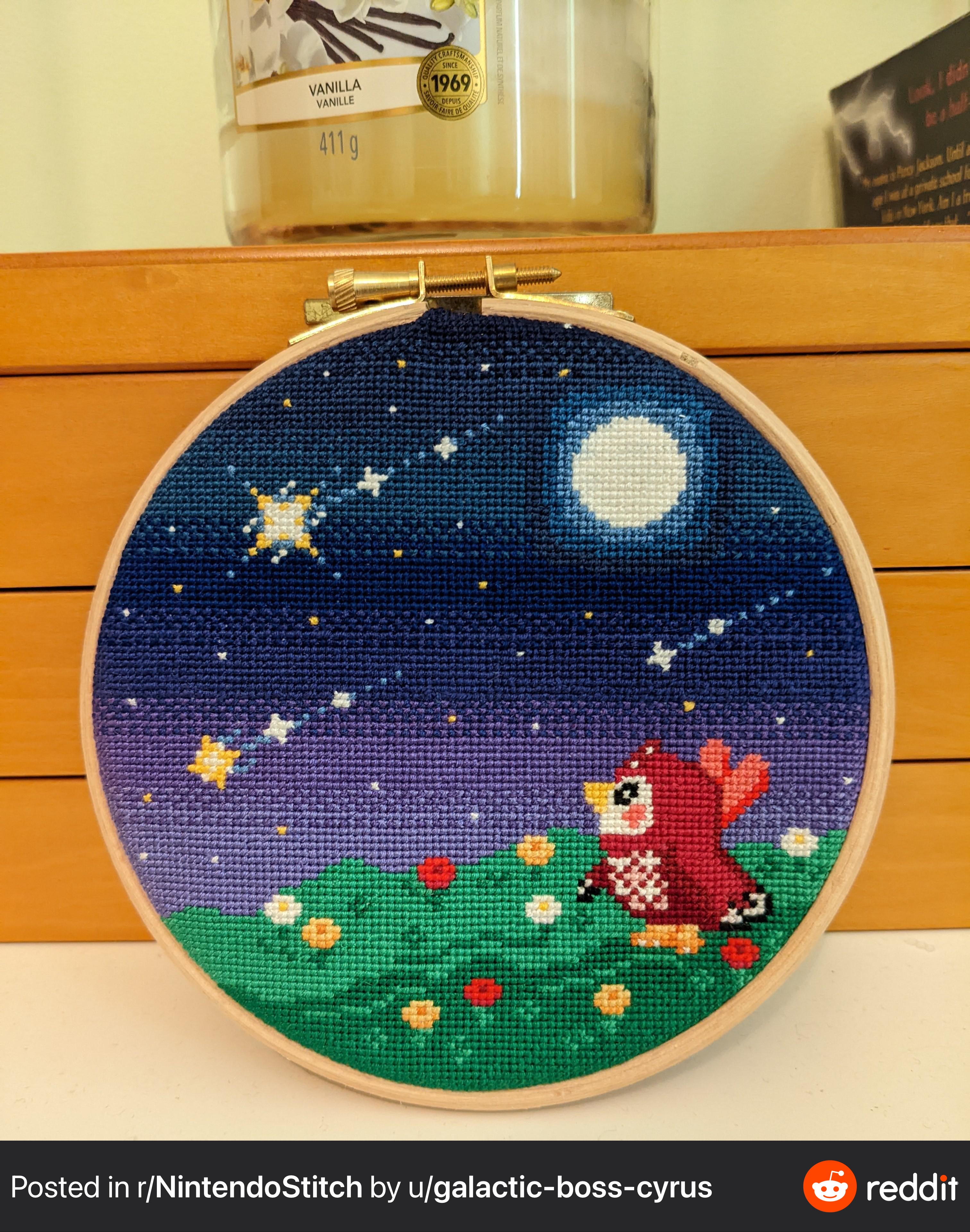 a closeup image of a crossstitch embroidery depicting a red bird from animal crossing: new horizons standing in a field of grass with flowers under a starry night sky with a moon. the embroidery is in a wooden hoop and is sitting on a wooden shelf. there is a glass jar with the label "vanilla" and the year 1969 visible in the background.