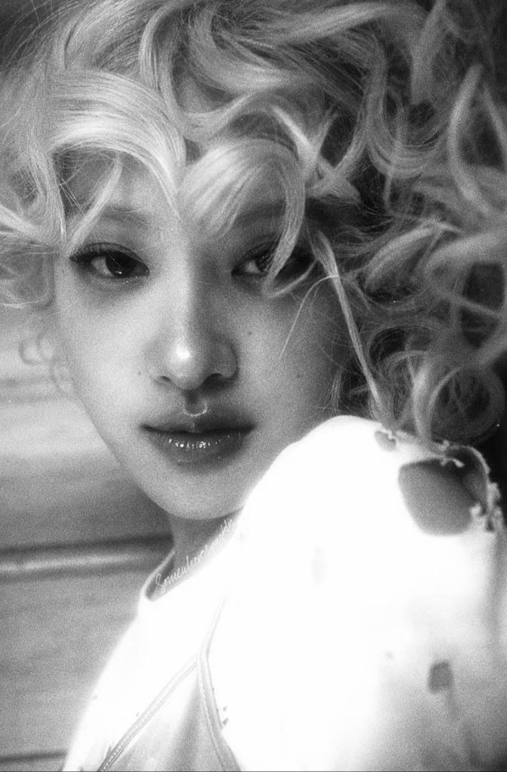 a black and white closeup portrait of a young woman with blonde, curly hair.  her face is partially obscured by her hair, creating a somewhat mysterious and sensual mood. she is wearing a lightcolored garment that covers her shoulders. the overall style is artistic and elegant, with a soft and slightly grainy texture suggesting a film photograph. the lighting is soft and focuses on her face and hair. the image evokes a sense of beauty and sophistication.