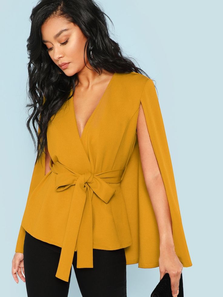a woman with long dark hair is modeling a mustard yellow cape top. the top has a vneck, a fitted waist with a selftie belt, and long sleeves with slits at the shoulder. she is wearing black pants and holding a black clutch. the background is a light blue.
