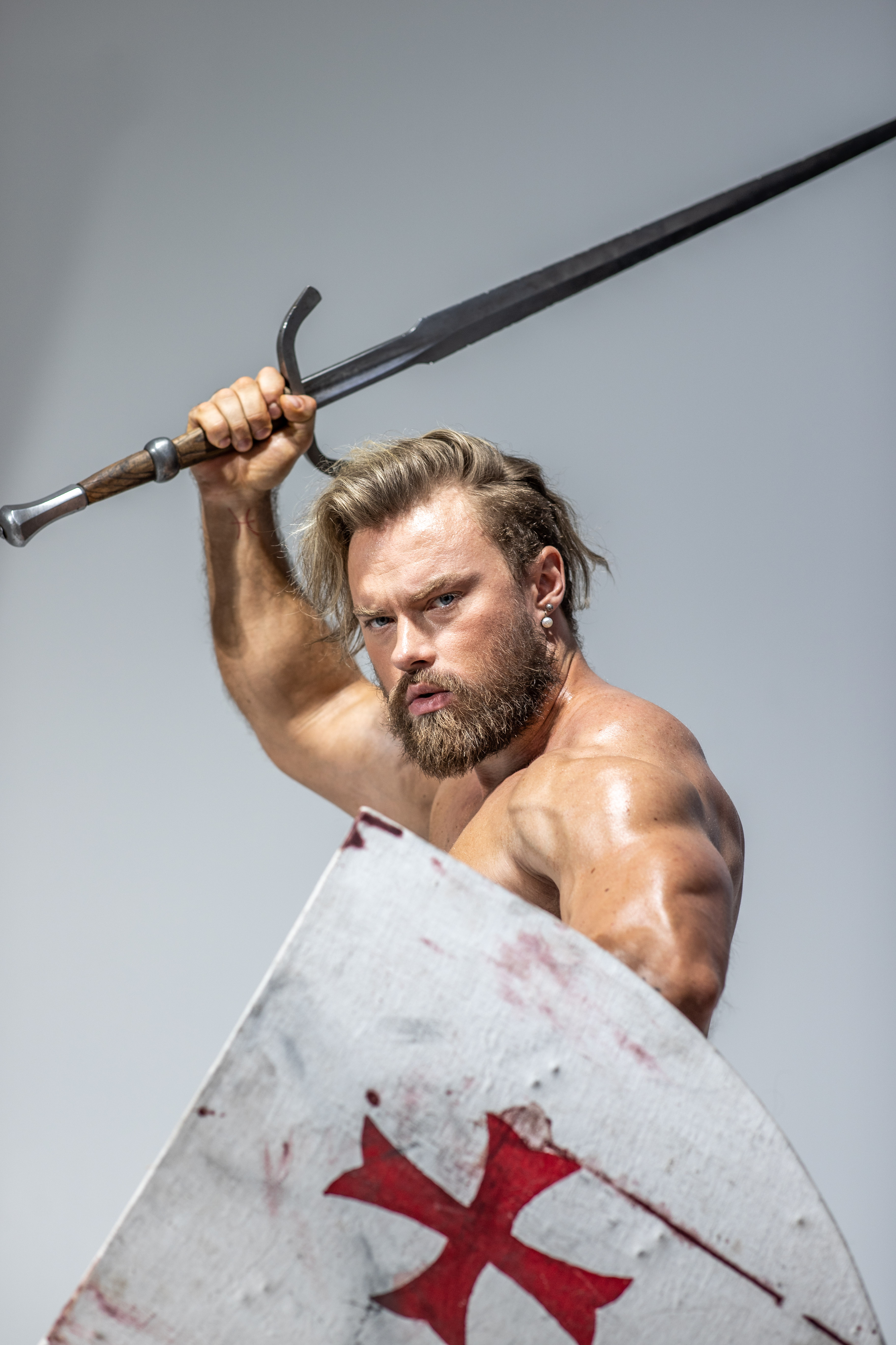 a muscular, bearded man in a dynamic pose, wielding a sword and shield. he appears to be a knight or warrior, possibly a templar, given the red cross on his shield. the image is taken from a low angle, emphasizing his strength and power. the studio setting provides a neutral backdrop, focusing attention on the subject. the lighting is bright, highlighting the details of his physique and the weaponry.