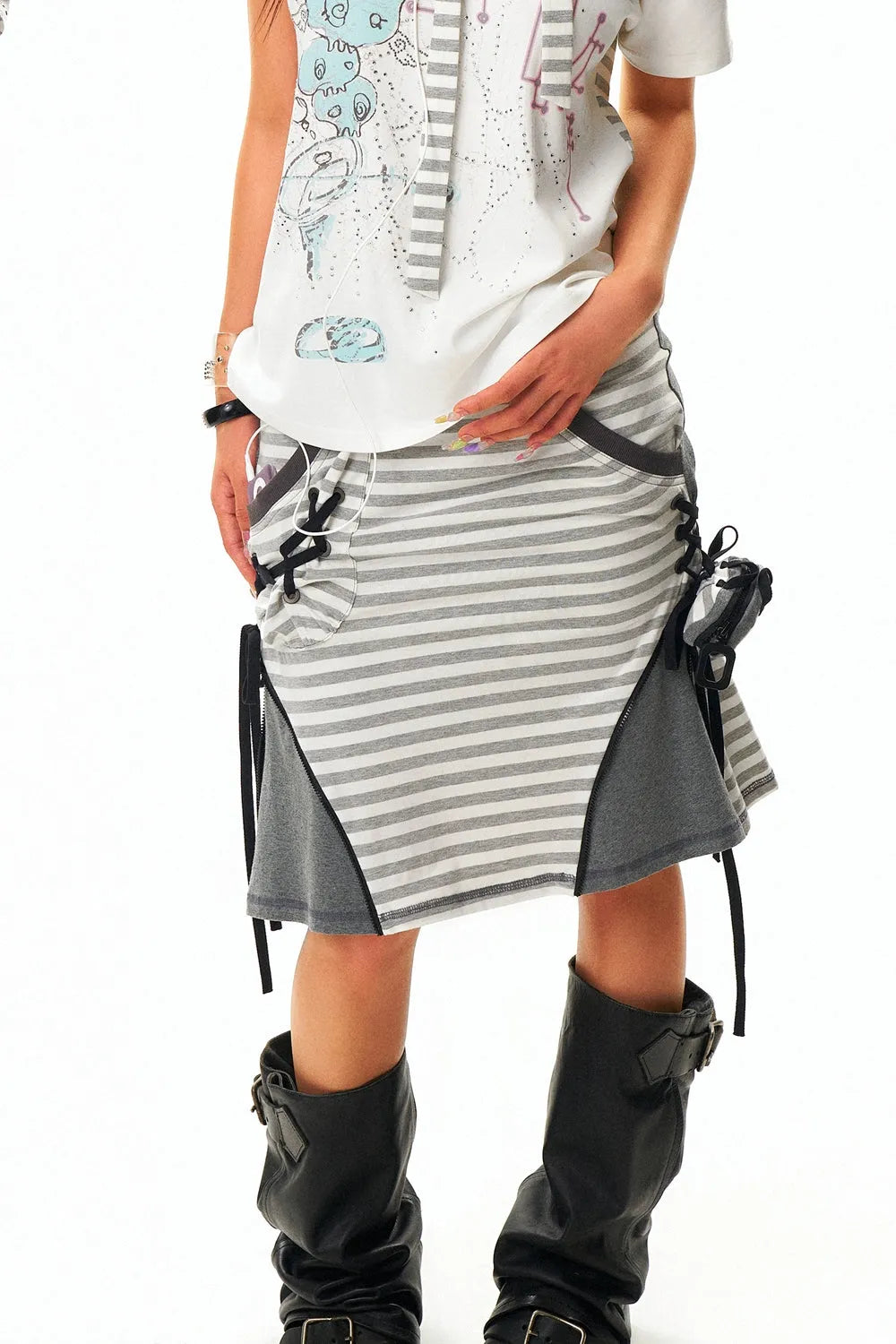 closeup shot of a young woman showcasing a stylish outfit. she is wearing a white and grey striped skirt with grey panels and black laces along the sides. the skirt has a slightly flared shape and is paired with a white, printed sleeveless top.  she is also wearing high black boots that have buckles. the woman's hands are visible, one resting on her hip and the other slightly visible near her side. the overall aesthetic is casual and fashionable. the background is plain white, focusing all attention on the clothing and model.