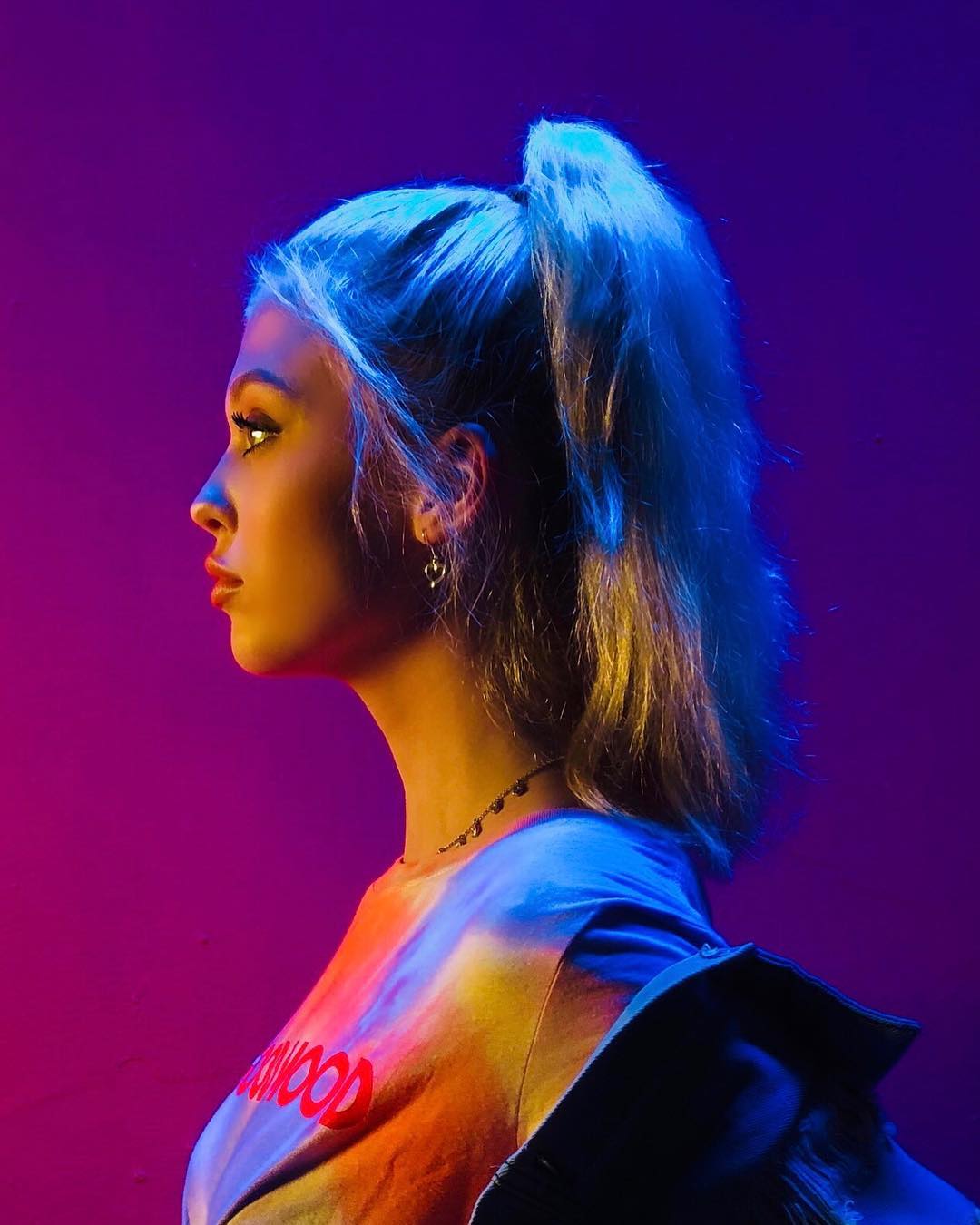 a profile shot of a woman with blue hair in a high ponytail, wearing a tiedye tshirt with red lettering, a necklace, and earrings. she has red lipstick and is posed against a pink background with blue and yellow lighting. a jacket is draped over her shoulder.