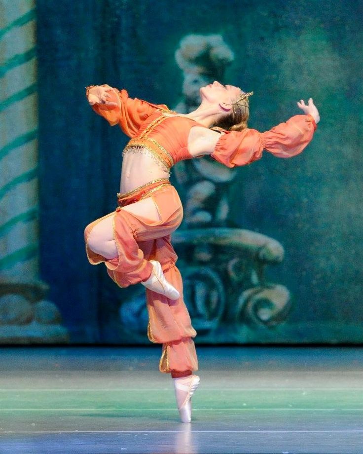 a ballerina in an orange, flowing costume performs an arabesque on pointe. she is elegantly poised, showcasing her grace and skill. the background features a painted backdrop suggestive of a theatrical set. the overall impression is one of beauty, grace and artistry. the costume is reminiscent of middle eastern or persian styles.