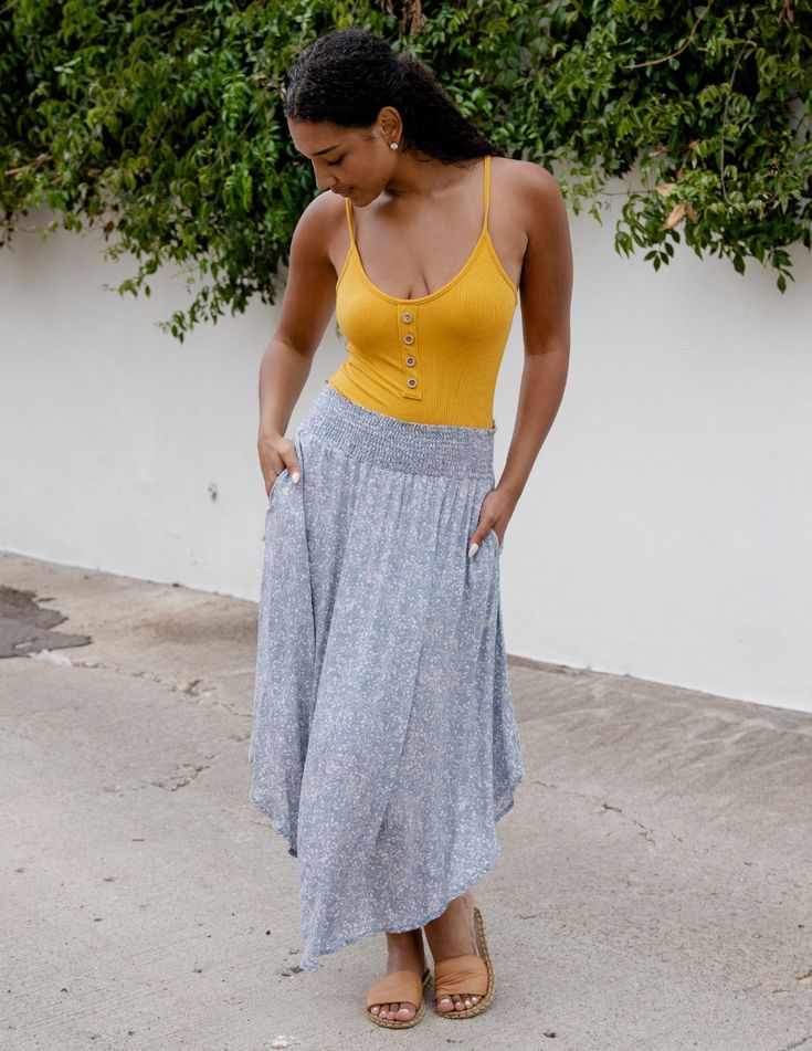 a young woman is standing on a sidewalk, modeling a casual summer outfit. she is wearing a yellow tank top and a long, flowing blue floral skirt.  she is also wearing brown sandals. the background is a white wall with some greenery.
