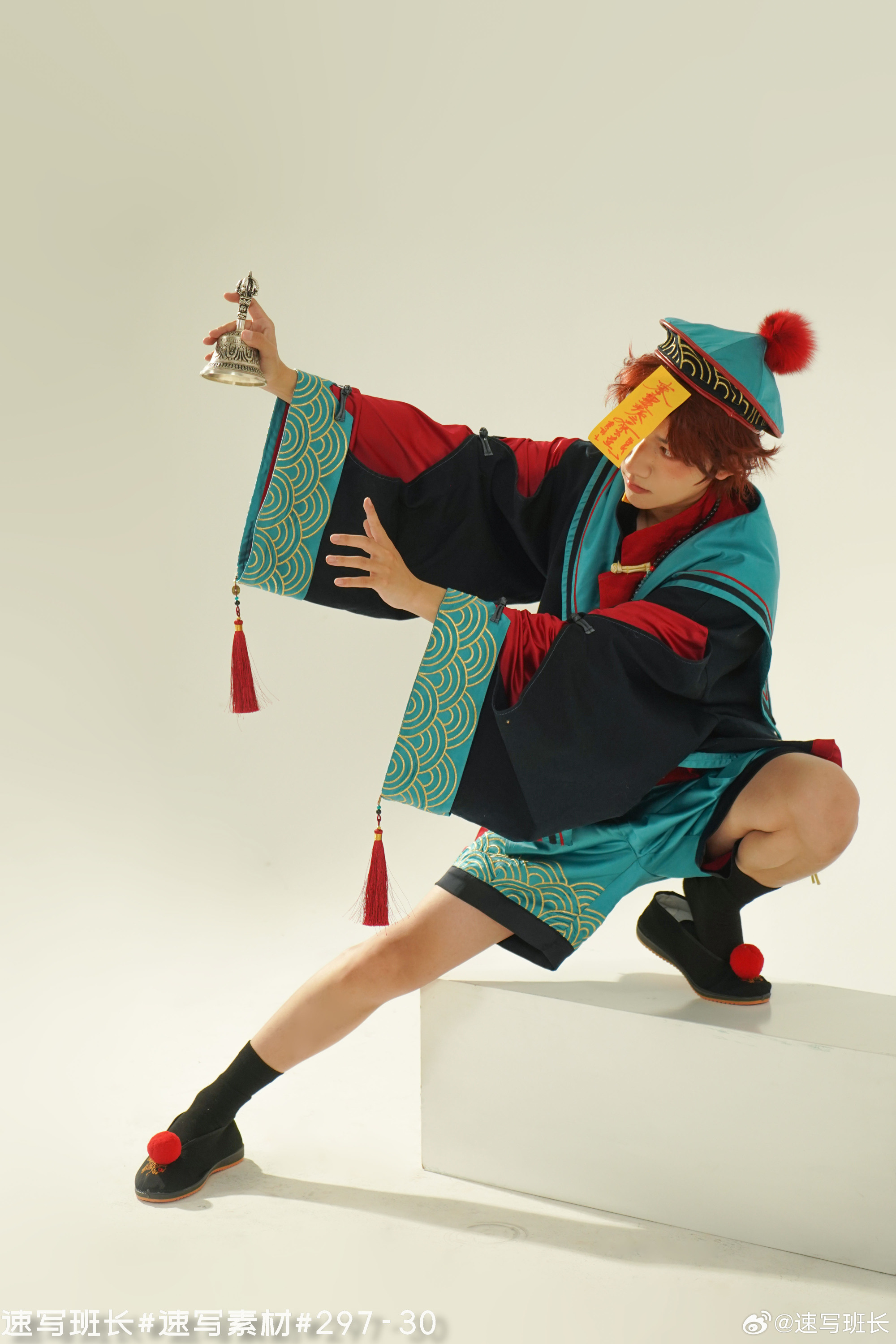a man in a vibrant teal and black chinese traditional costume with red accents poses dynamically on a white cube. he holds a small silver bell in his right hand and a yellow paper talisman is stuck to his forehead.  he wears black socks and black cloth shoes with red pompoms.  the scene is brightly lit in a studio setting, captured from a low angle that emphasizes his pose.