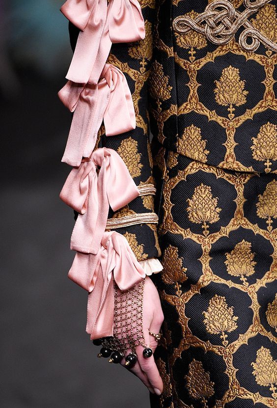 a closeup shot focusing on the sleeve and hand of a model showcasing a gucci outfit. the sleeve is adorned with multiple layers of delicate pink satin bows.  the fabric of the jacket and skirt is a rich black brocade with a gold, ornate pattern. the model's hand wears a chainlink bracelet with dark beads, and several rings. the image highlights the intricate details and luxurious textures of the clothing and accessories. the overall aesthetic is one of high fashion and elegance.
