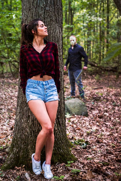 a still image from a horror film depicting a young woman in distressed denim shorts and a red plaid shirt standing against a tree in a forest. she appears frightened. a masked man with a machete is visible in the background, suggesting he is stalking or pursuing her. the scene is set in a forest with autumn leaves on the ground. the overall mood is suspenseful and terrifying.
