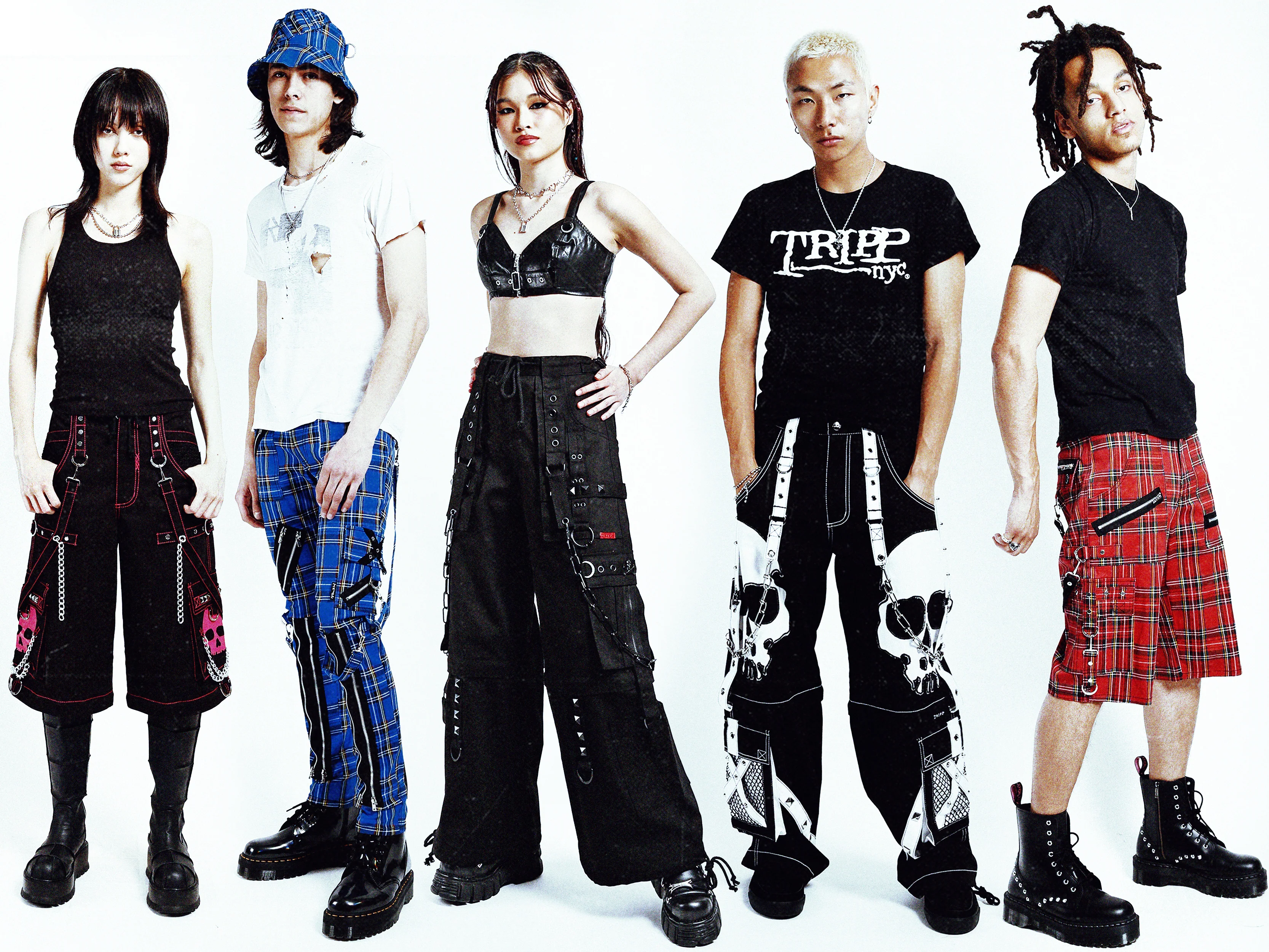 studio shot against a white background shows five young models, three female and two male, dressed in punk/gothinspired clothing. the clothing is characterized by dark colors, plaid patterns, cargo pants, harness details, and skull motifs.  each model has a unique style, showcasing different pieces of clothing but a cohesive theme. the models' poses and expressions are cool and confident.  the overall impression is one of alternative fashion and youth culture. one of the male models is wearing a tripp nyc tshirt.