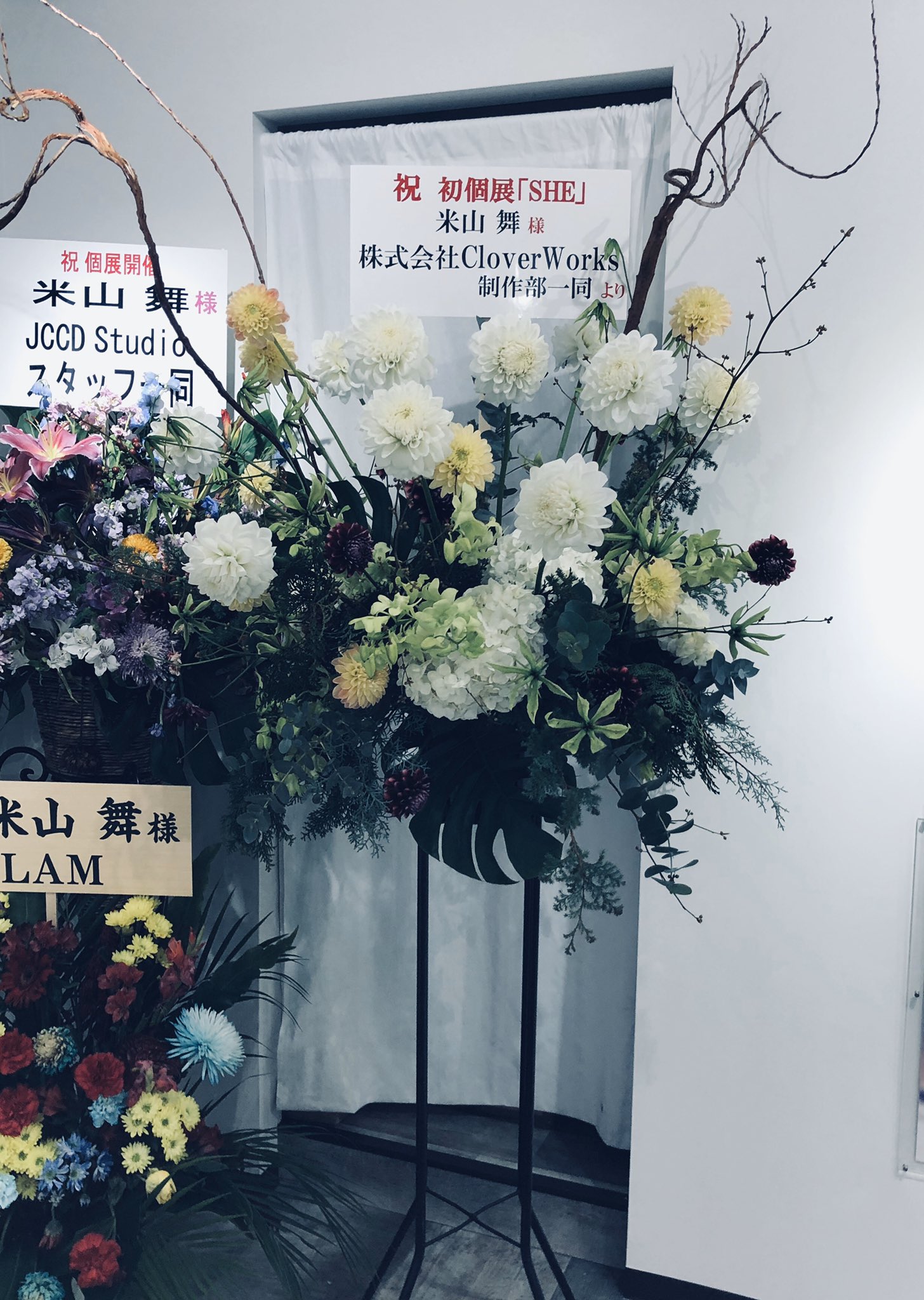 a photo of a flower arrangement with a congratulatory message for an artist's first solo exhibition.  the arrangement is placed in front of a white curtain, with a sign on each side. the sign on the left reads "congratulations on the solo exhibition. to ms. mai yoneyama, from all jccd studio staff." the sign in the middle reads "congratulations on your first solo exhibition "she", ms. mai yoneyama, from clover works production department. " the sign on the right reads "ms. mai yoneyama, lam".  the flower arrangement consists of white and light green flowers.