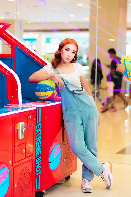 a young redhead woman in light blue denim overalls leans against a red and blue arcade basketball game machine. she is holding a small basketball and smiling slightly. the setting appears to be a brightly lit shopping mall with other people visible in the blurred background. the arcade game is prominently featured in the foreground. the overall mood is fun and playful.