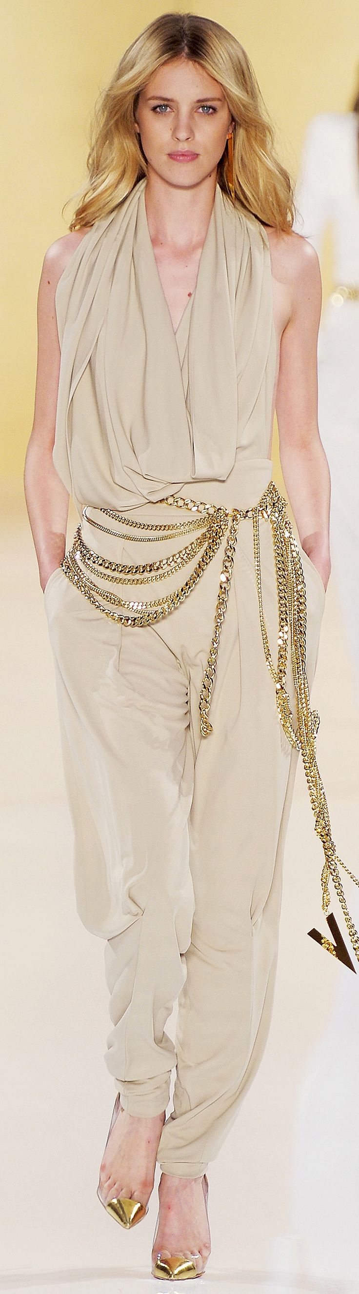 a fashion model walks the runway in a white dress with a gold chain belt. she is wearing her hair down and has a neutral makeup look. the model is posing for the camera.