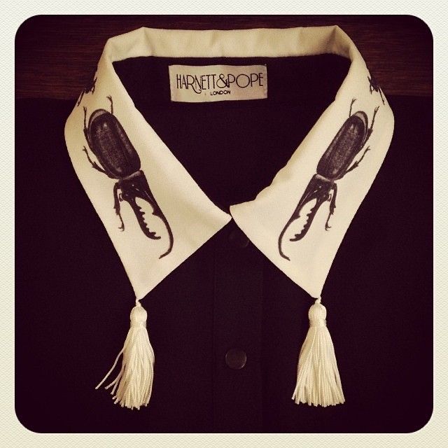 a black shirt with a white collar, featuring beetle designs and tassels. the collar has a tag that reads "harnett & pope london".