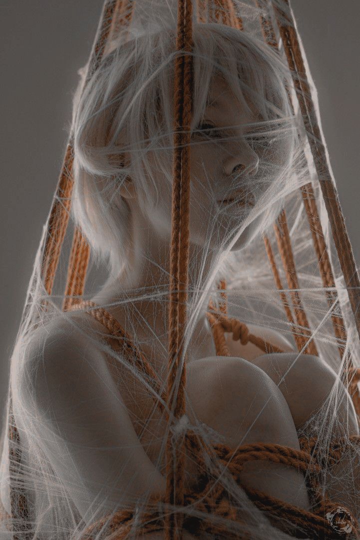 he image shows a closeup portrait of a blonde woman with pale skin. she is enveloped in a delicate spiderweblike structure made of thin white threads and thicker orange ropes. the ropes appear to be binding her, suggesting a sense of entrapment. the woman's expression is partially obscured by the web, adding to the mysterious and ethereal mood. the overall aesthetic is artistic and conceptual, with a blend of fantasy and sensuality.  the lighting is soft and diffuse, creating a subtle contrast between light and shadow. the background is a simple, neutral gray, keeping the focus on the subject and the intricate texture of the web and ropes.