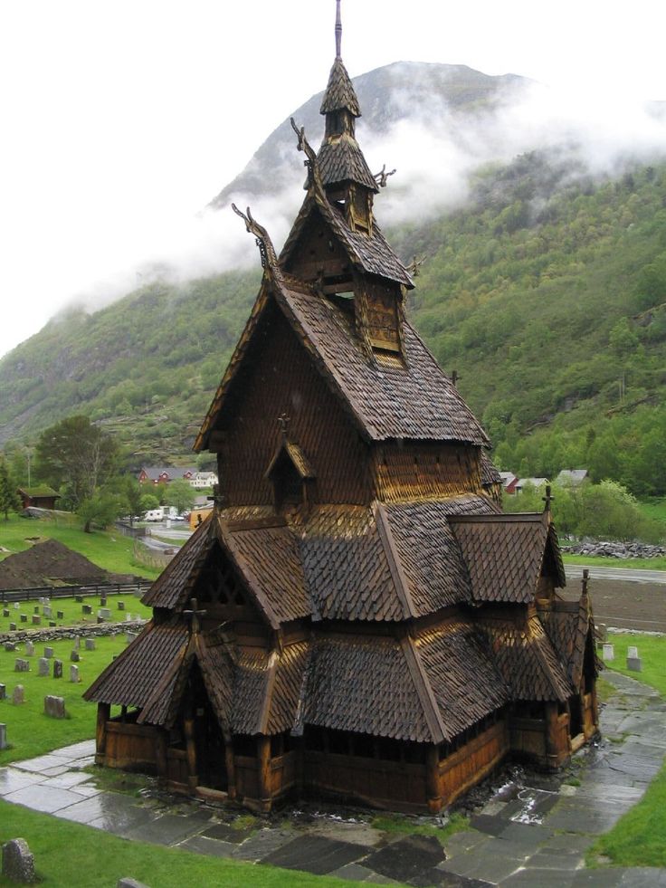the image showcases a beautifully preserved urnes stave church, a prominent example of traditional norwegian timber architecture. the church's multitiered roof covered in dark shingles and intricate wooden carvings is the central focus. the structure stands in a graveyard, with simple headstones scattered across a grassy field. behind the church, a lush green mountain shrouded in mist adds to the picturesque landscape. the setting suggests a rural location and a site of historical and cultural significance. the building is a significant example of stave construction and is a popular tourist destination. the day seems to be overcast, contributing to a serene, somewhat mystical atmosphere.