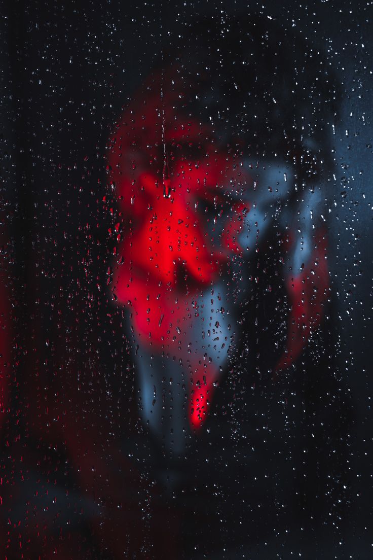 the image is an abstract and moody portrait seemingly taken through a raincovered window at night. the subject's face is blurry and obscured by the water droplets on the glass, making the identity and expression indistinct. red and blue neon lighting creates a dramatic contrast against the dark background. the overall effect is atmospheric and mysterious, lending itself to interpretations of loneliness, isolation, or hidden identity. the focus is not on sharp detail but rather the feeling and texture of the scene, heavily relying on color, light, and the blur created by the water droplets.