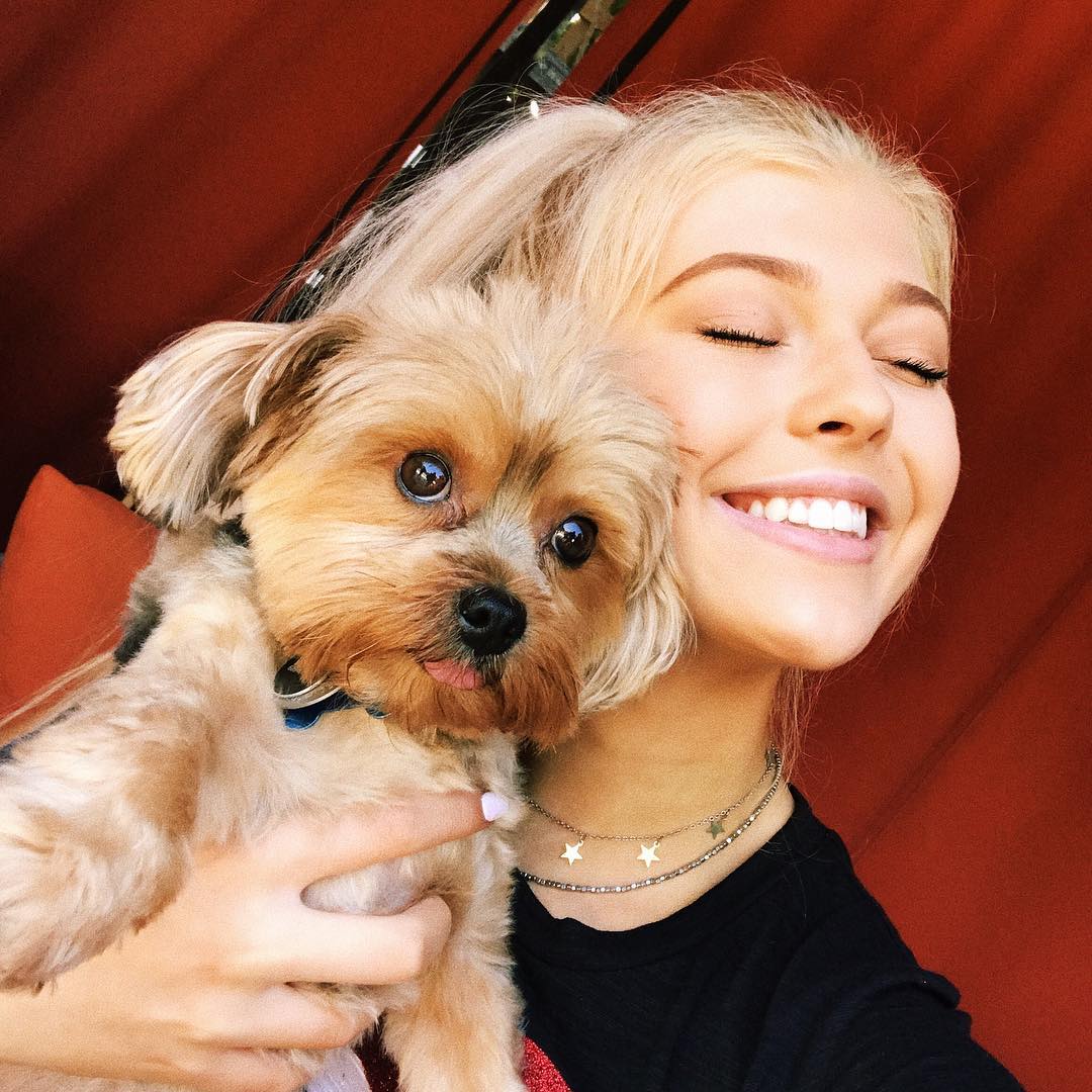 a blonde woman with her eyes closed and a big smile on her face, hugging a brown furry dog with its tongue slightly sticking out. they are in front of a red background. the woman is wearing a star necklace and a black shirt.