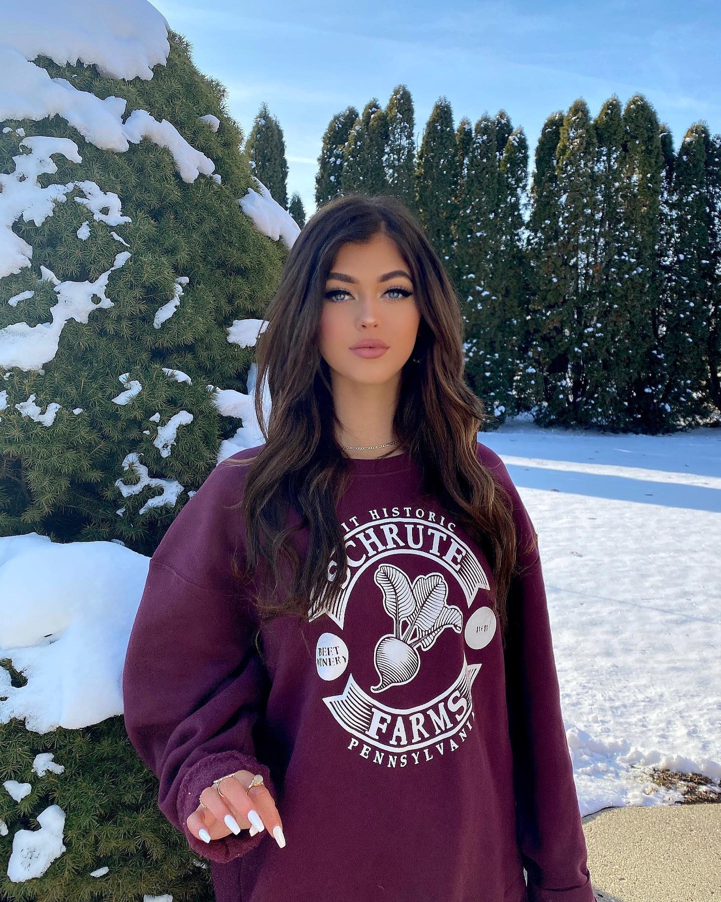 a woman is standing outside in the winter wearing a schhrute farms sweatshirt. she is surrounded by snowcovered trees.