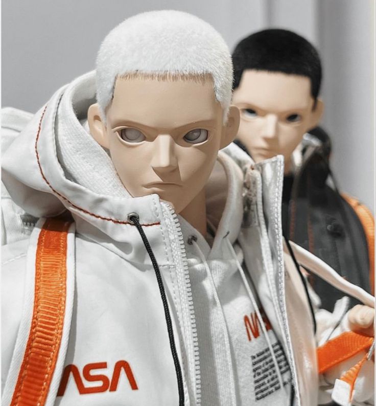 a close up of a white male doll wearing a white astronaut jacket. the doll is standing and looking at the camera. the doll is wearing a black zipper and an orange zipper. the doll's right arm is bent and the left arm is straight down. behind the doll, the viewer can see a second doll wearing a black jacket.