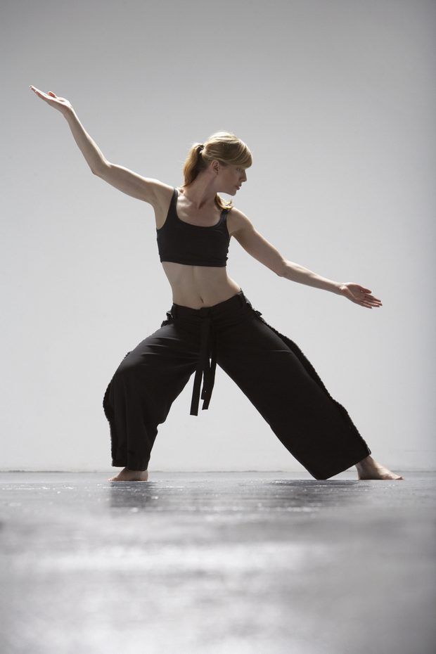a woman wearing black pants and a black top is stretching and doing a karate pose, with her left leg forward and right leg back, her arms are stretched out to the sides and she is looking down. the background is grey.