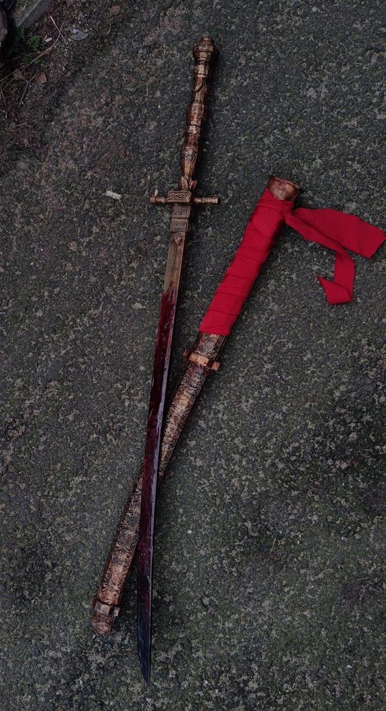 two prop swords lay crossed on a dark asphalt ground. one sword is longer with an ornate and detailed handle. the other, shorter sword is wrapped in red fabric. both appear to be covered in a bloodlike substance.