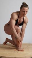 a muscular woman kneels on her right knee on a round wooden platform. she is wearing a black bikini bottom and a black sports bra. she is lit with soft studio lighting and is posed in a way that shows off her physique.