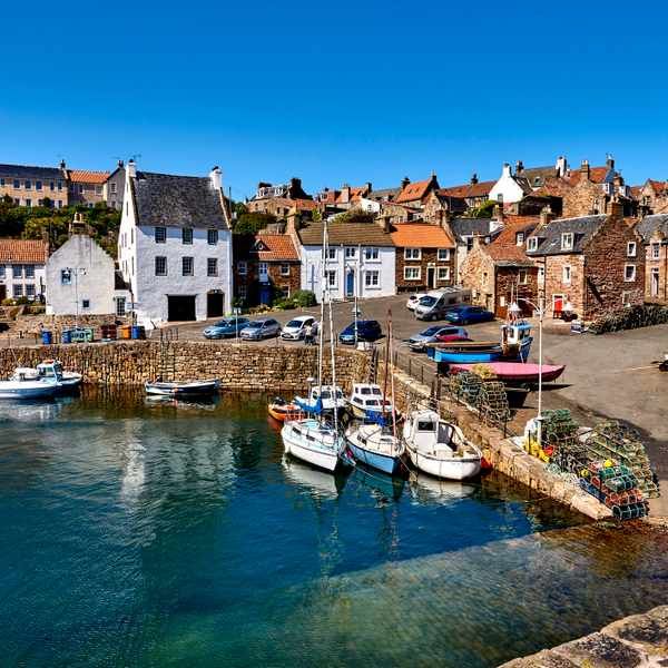 the image showcases a picturesque fishing village, likely in scotland, on a bright sunny day.  the harbor is filled with a variety of small fishing boats and sailboats, neatly moored along the stone harbor wall.  the village itself is charming, with tightly packed stone houses in traditional architectural styles. cars are parked in a small lot overlooking the harbor. the water in the harbor is calm and reflects the blue sky and colorful boats. the overall scene is idyllic and peaceful, suggesting a popular tourist destination.