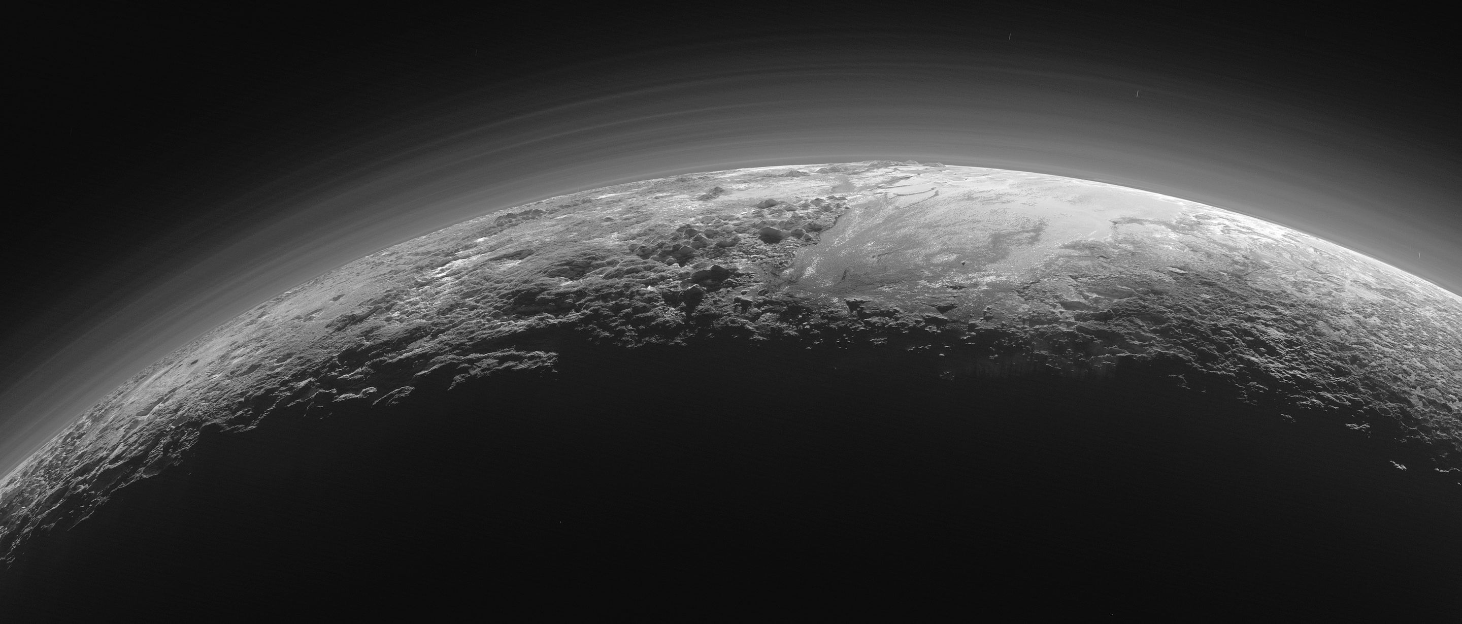 this is a black and white image of pluto, a dwarf planet located in the kuiper belt beyond neptune. the image shows the crescent of pluto, with the sunlit side of the planet facing the camera. the surface of pluto is covered in a mix of ice and rock, and a thin atmosphere can be seen as a faint haze surrounding the planet.  the image is taken from a distance, with the majority of the image being the black of space with a few faint stars visible.