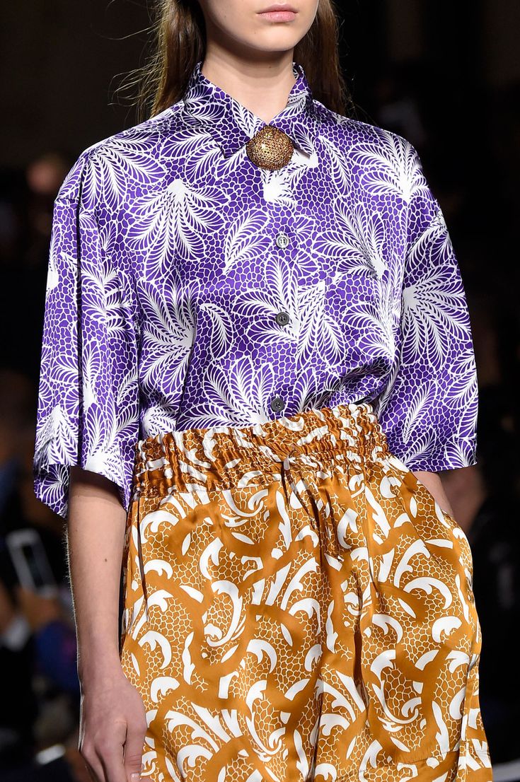 closeup shot of a fashion model showcasing a striking outfit on a runway. the model is wearing a purple, shortsleeved shirt with a white leaflike pattern, paired with mustardyellow highwaisted shorts featuring a similar intricate design. a gold brooch adds a touch of elegance to the collar of the shirt. the overall aesthetic is rich in texture and pattern, suggestive of an autumn/winter collection. the focus is on the clothing's intricate details and the luxurious fabrics. the backdrop is blurred, keeping attention on the model and the outfit.
