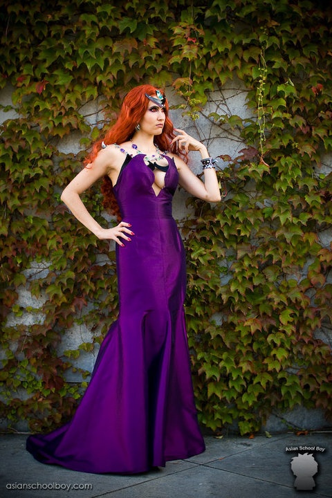 a woman dressed in a purple gown with jewelry and a headpiece stands in front of a wall covered in ivy. she is posing for the camera.