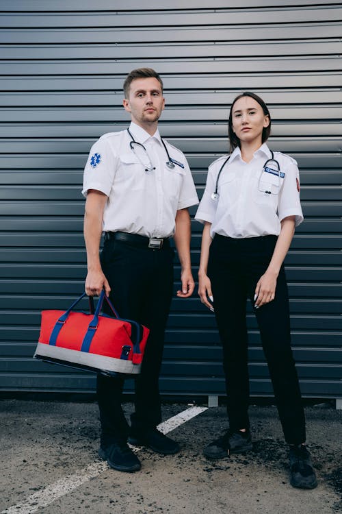 two paramedics, a man and a woman, stand sidebyside in front of a grey metal wall. they are both wearing white shortsleeved uniforms with black pants and stethoscopes around their necks. the man holds a red medical bag. they are looking straight ahead with serious expressions. the image conveys a sense of professionalism and teamwork.
