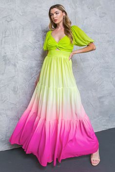a young woman in a stylish maxi dress stands against a plain wall. the dress is a beautiful ombre design, fading from green to yellow and then to pink. the bodice is a fitted, sleeveless top with a twist detail in the center. the skirt is flowy and tiered, creating a whimsical look. she is wearing a pair of white heels and looking at the camera with a confident smile.