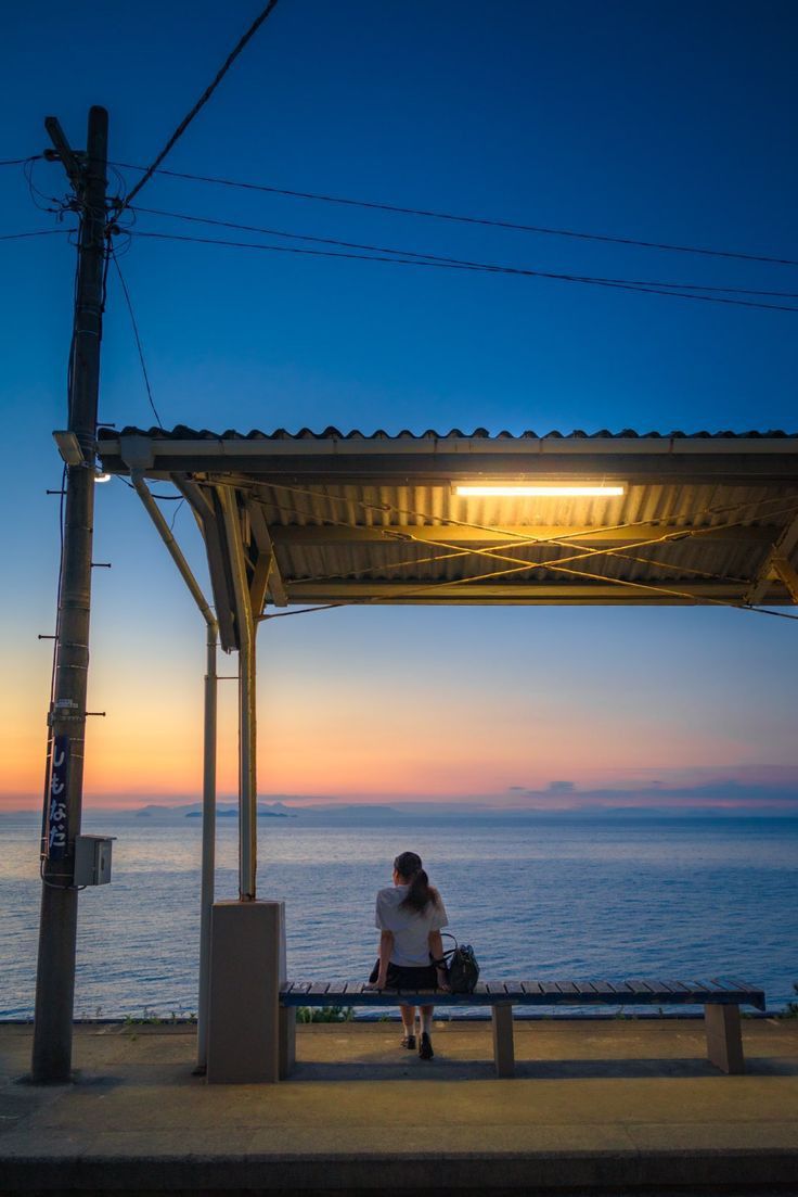 a young woman sits alone on a bench at a small, seaside train station, watching the sunset over a calm ocean. the sky is a beautiful blend of oranges, pinks, and blues. the station shelter is lit by a single fluorescent light. the scene conveys a sense of peace and solitude. the overall feeling is one of tranquility and quiet contemplation.