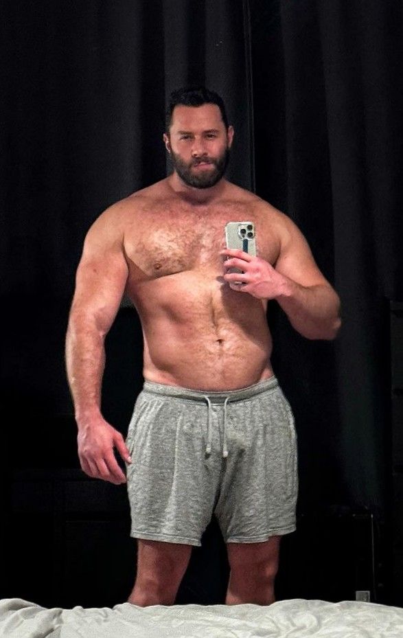 muscular, hairychested man takes a selfie in his bedroom. he is wearing gray shorts and is standing in front of a dark background. he appears to be in his 30s or 40s and has a wellbuilt physique.  the image is a fulllength shot from the waist up.  the man is holding his iphone to take the picture.