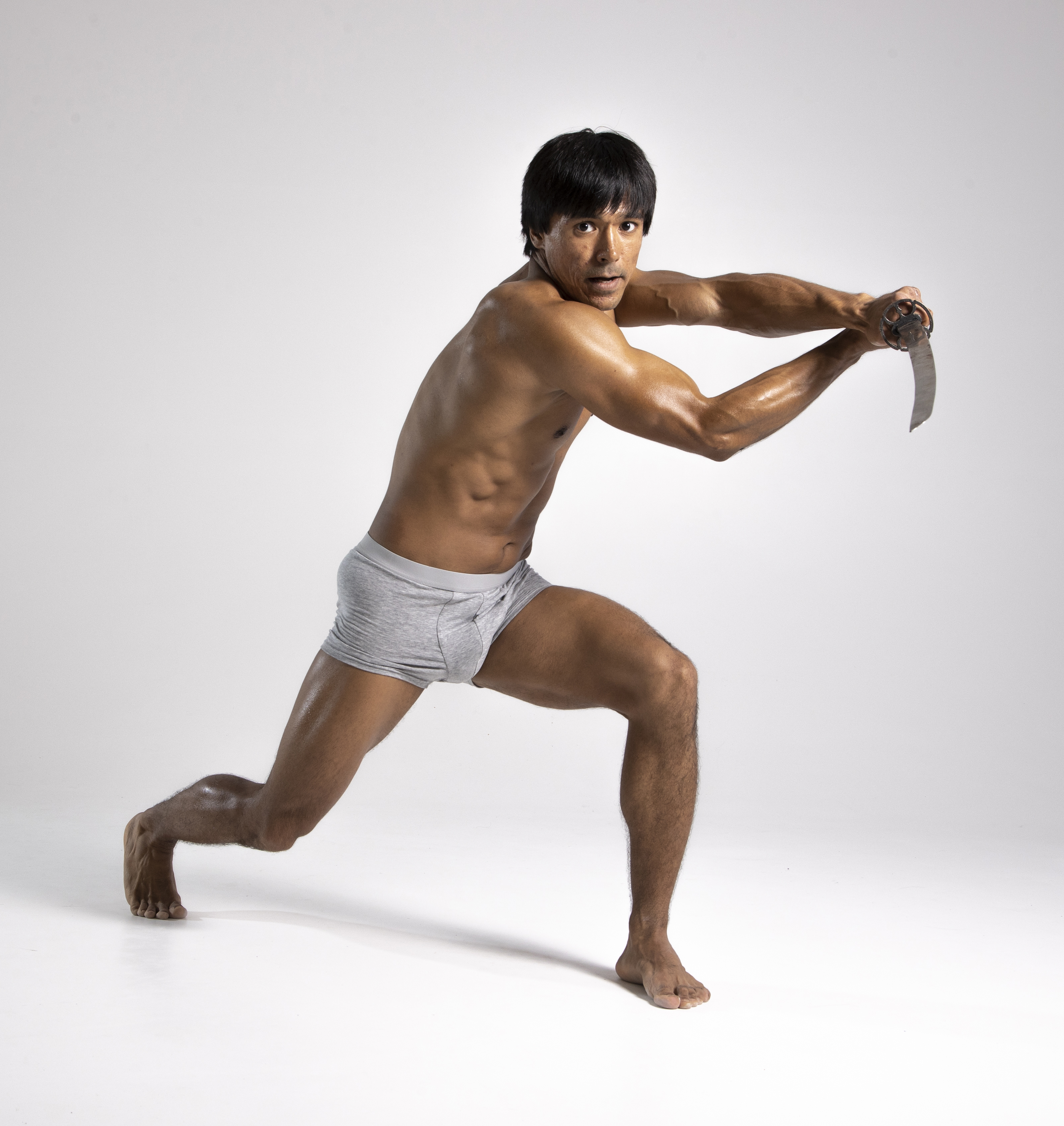 a muscular man in gray underwear is barefoot and in a martial arts stance with a sword. he is set against a white background.