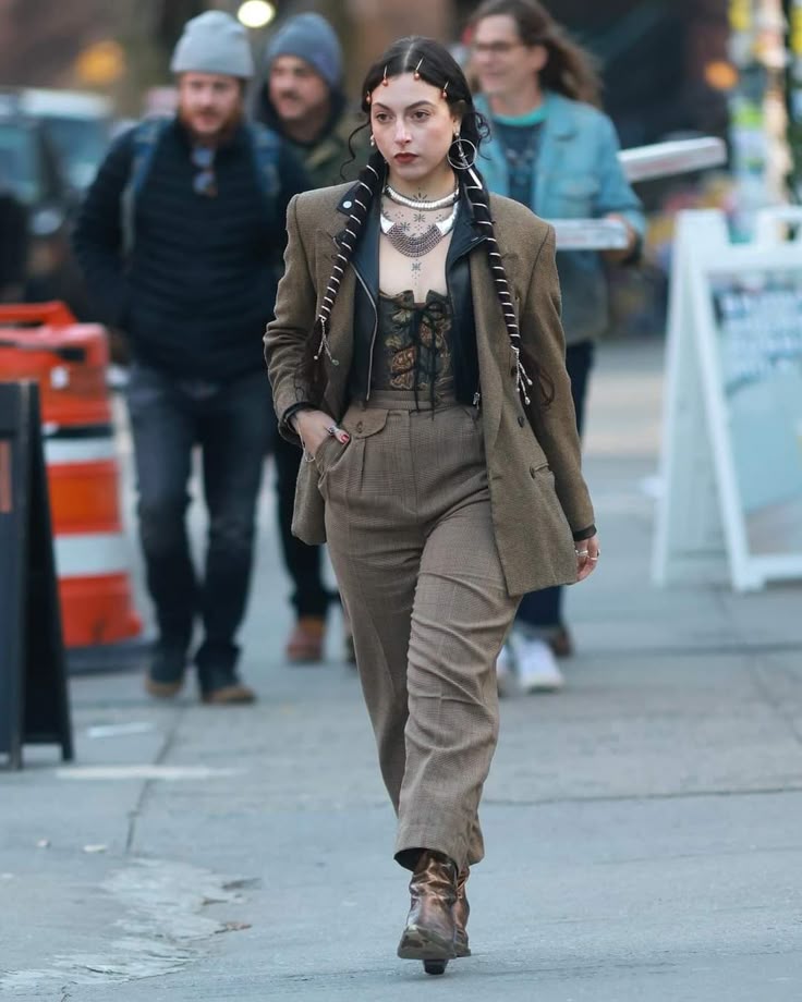 a woman in a blazer, pants, and boots walks on a city street. she has braids, a tattoo, and a necklace. she is wearing a corset and a black top under her blazer. there are other people in the background.