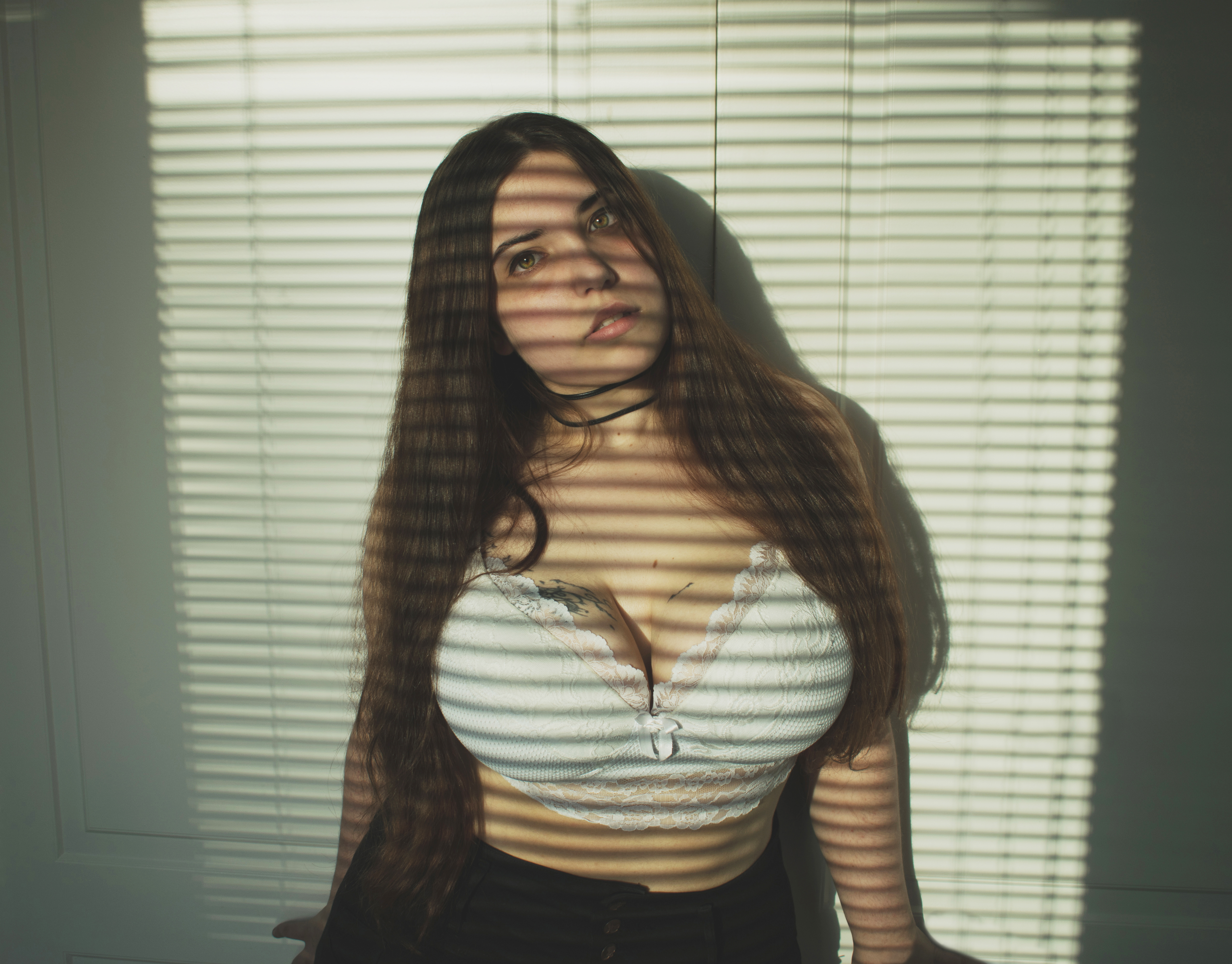 a woman with long brown hair is standing in front of a wall with blinds, casting shadows across her face and body. she is wearing a white top with lace trim and a black choker. a tattoo is visible on her chest.