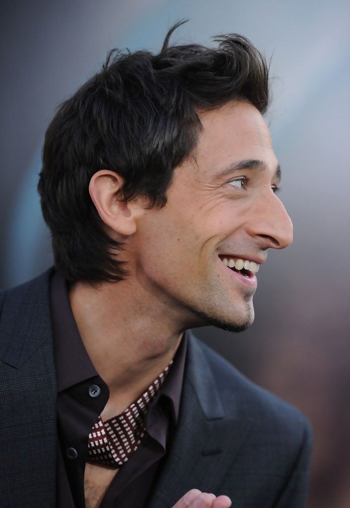 a closeup profile shot of a man, possibly an actor, smiling warmly. he's dressed in a darkcolored suit and shirt with a patterned tie. his dark hair is styled neatly. the background is blurred, suggesting an outdoor event or red carpet setting. the overall impression is one of a confident and stylish individual.