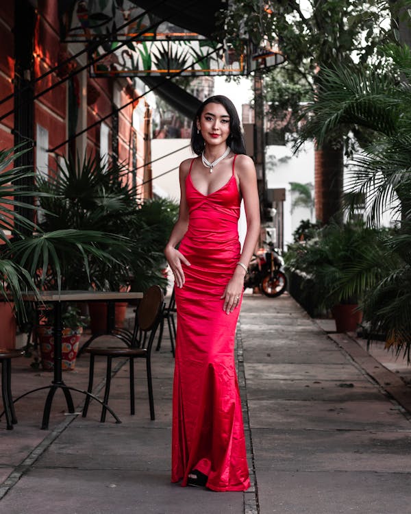 a fullbody shot of a young, attractive asian woman in a vibrant red satin maxi dress. she is standing confidently on a paved walkway between rows of lush tropical plants, with the architecture of a building visible in the background. the dress is a figurehugging, floorlength style with a deep vneck. she wears a delicate necklace and appears to be posing for a photograph. the overall ambiance is elegant and sophisticated.  the setting suggests an outdoor urban or possibly tropical location.