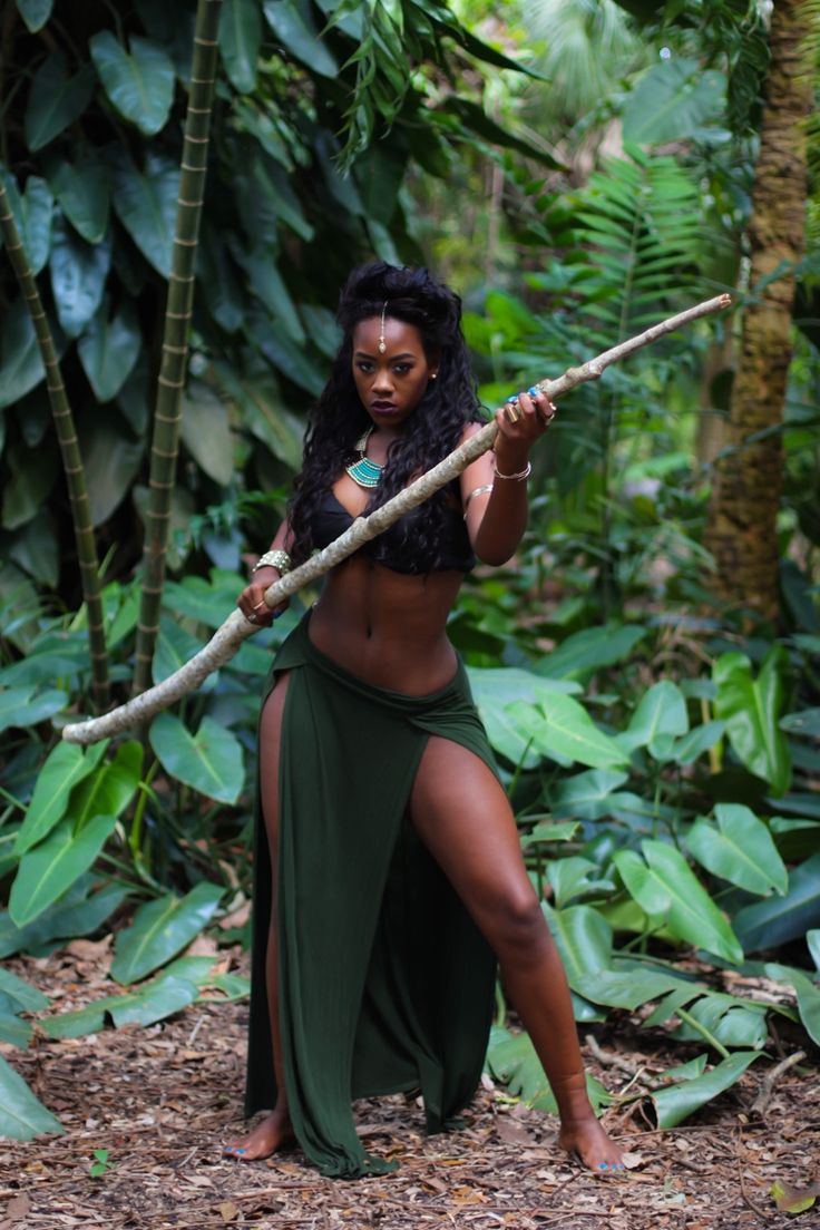 a darkskinned woman with long, dark hair poses confidently in a lush tropical rainforest setting.  she's wearing a dark green, thighhigh split skirt and a matching crop top.  she holds a long, sturdy branch like a staff or weapon. the overall mood is fierce, strong, and suggestive of a tribal warrior or amazonian goddess. the background is filled with vibrant green foliage, highlighting the contrast between the woman and her environment. the image is welllit and professionally photographed, exhibiting a sense of both power and natural beauty.