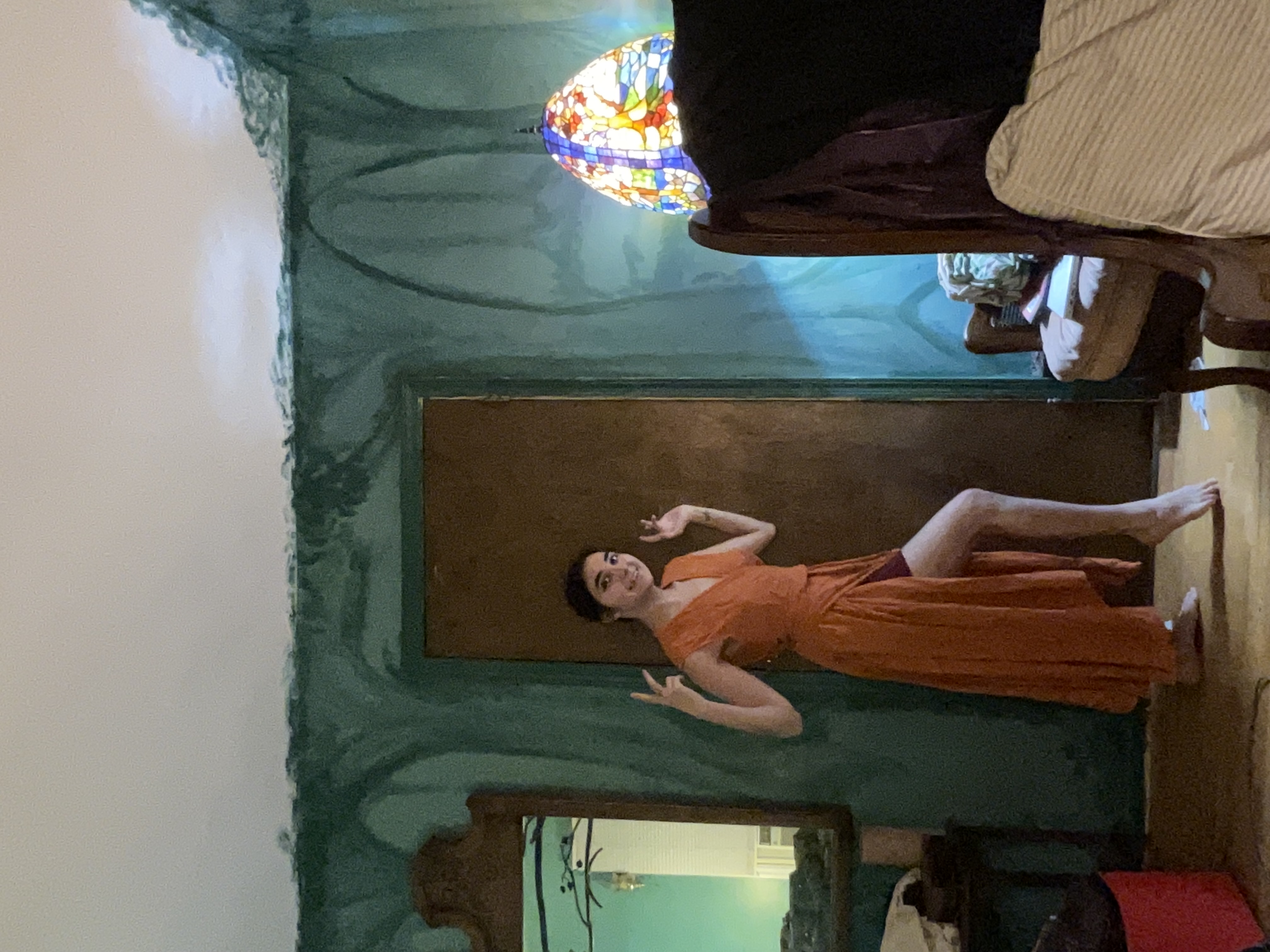 a young woman in a long orange wrap dress with a high slit stands in a bedroom with teal painted walls. she is posing, making a peace sign with one hand. the room has an armchair, a stained glass lamp, and a mirror.