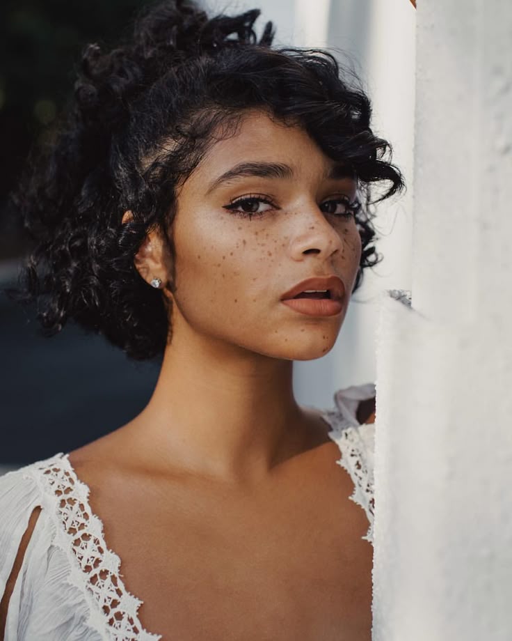a closeup portrait of a woman with curly dark hair, freckles, and wearing a white dress with lace trim. she is standing next to a white column and looking directly at the camera.